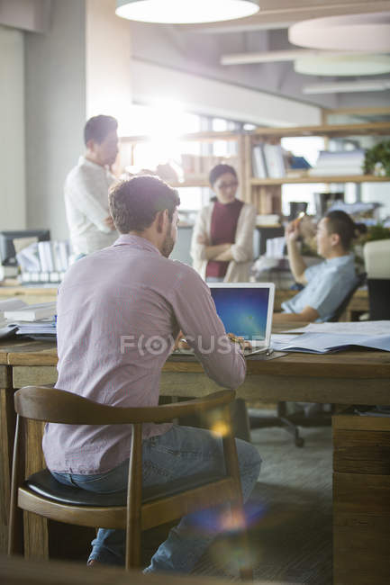 Вид спереду офісного працівника, який використовує ноутбук з колегами на задньому плані — стокове фото