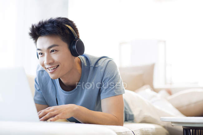 Chinois dans les écouteurs à l'aide d'un ordinateur portable sur le canapé — Photo de stock
