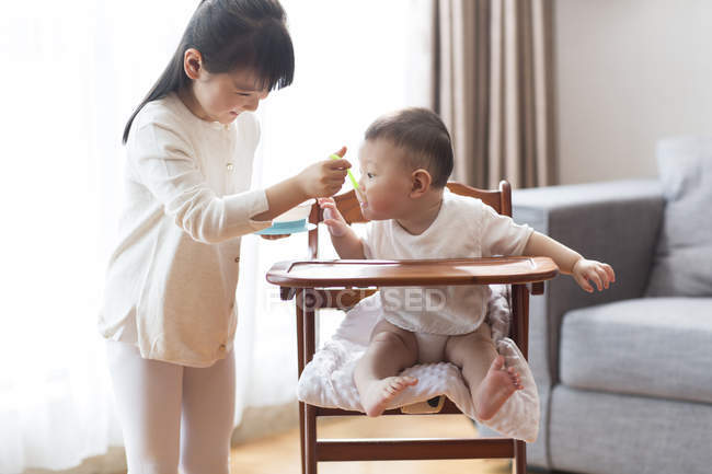 Niña china alimentando a bebé niño en silla alta - foto de stock