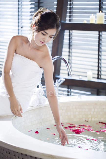 Young Chinese woman sitting by bathtub with rose petals — Stock Photo