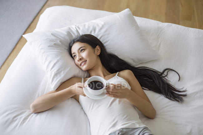 Chinesin liegt mit Tasse Kaffee im Bett — Stockfoto