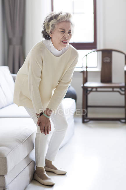 Senior chinesische Frau hält Knie in Schmerzen im Wohnzimmer — Stockfoto