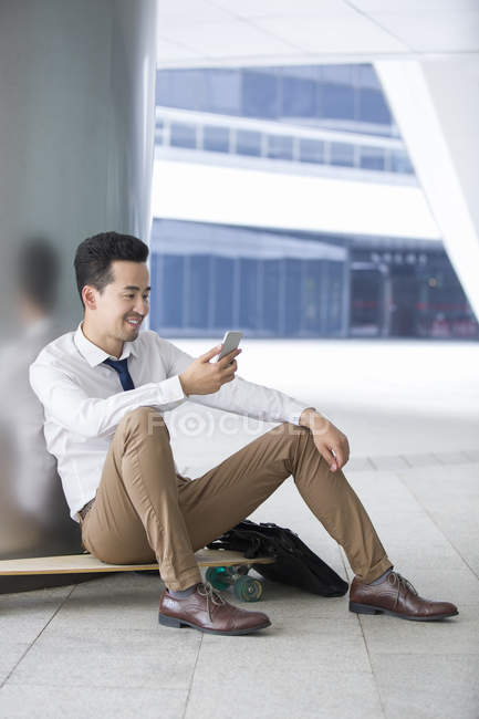 Азиатский бизнесмен использует смартфон, сидя на скейтборде на парковке — стоковое фото