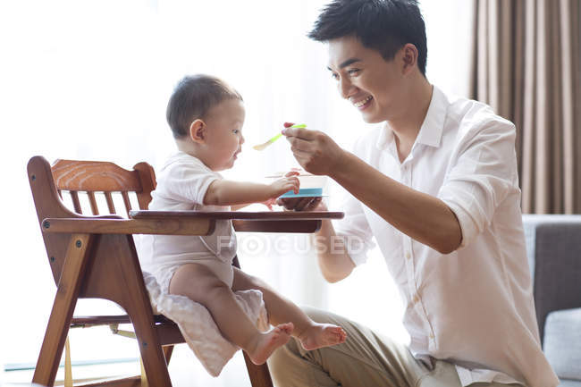 Chinese father feeding baby boy in high chair — Stock Photo