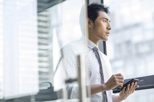 Chinesischer Geschäftsmann mit Smartphone im Büro und Blick durchs Fenster — Stockfoto