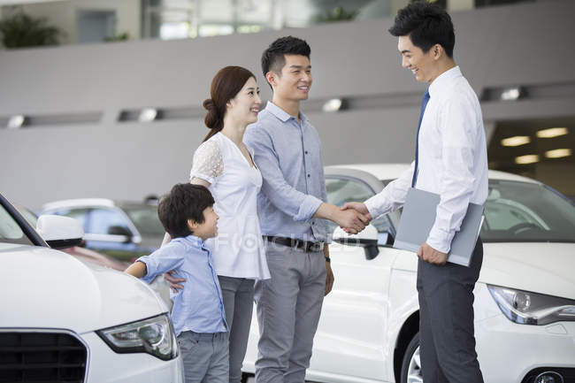 Familia china estrechando la mano con vendedor de coches en sala de exposición - foto de stock