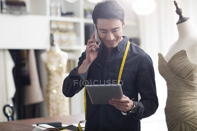 Diseñador de moda asiático hablando por teléfono en el estudio y la celebración de la tableta digital - foto de stock