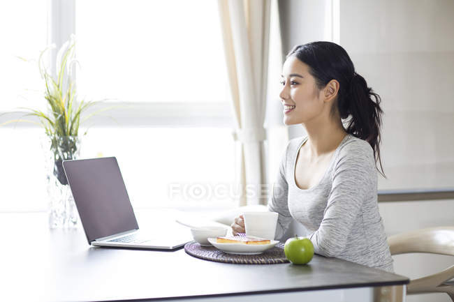 Mujer asiática usando el ordenador portátil mientras desayuna - foto de stock