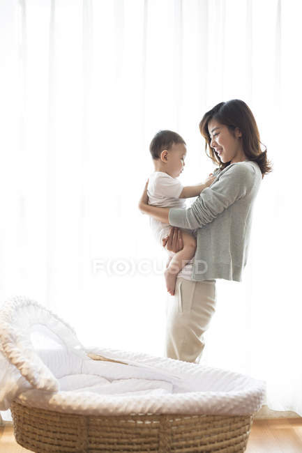 Madre china sosteniendo bebé niño en la habitación con cuna - foto de stock