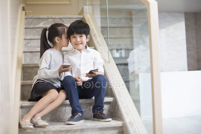 Chinese boy and girl holding smartphones and whispering on stairs — Stock Photo