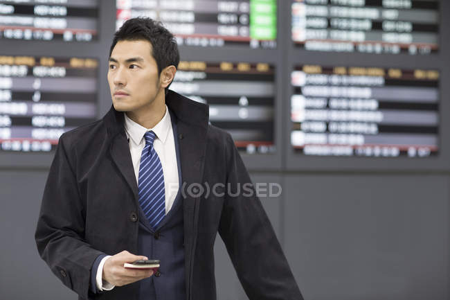 Hombre de negocios chino esperando en el aeropuerto con pasaporte y teléfono inteligente - foto de stock