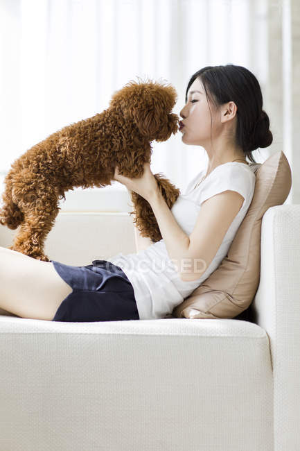 Joven mujer china jugando con caniche mascota en el sofá - foto de stock
