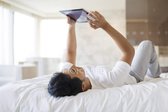 Chinese man lying down with digital tablet on bed — Stock Photo