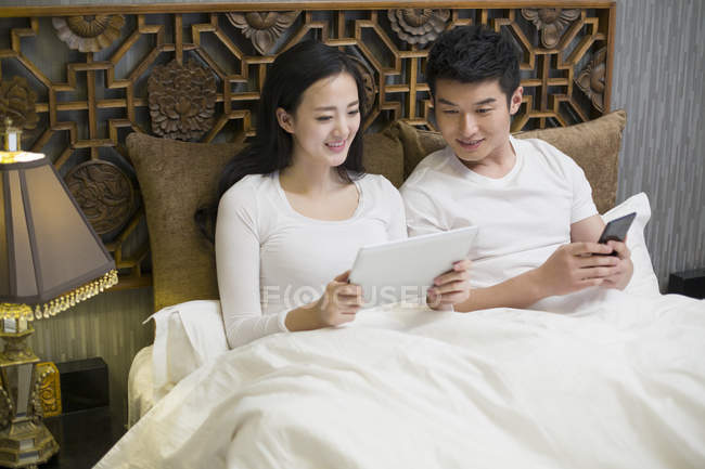 Casal asiático usando tablet digital e smartphone na cama — Fotografia de Stock