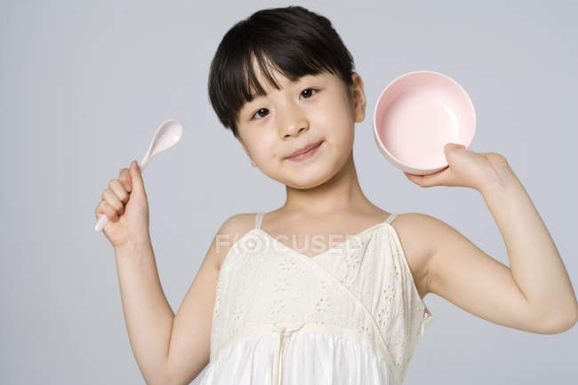 Niña china sosteniendo tazón y cuchara sobre fondo gris - foto de stock