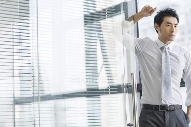 Chinese businessman leaning on doorway in office and looking away — Stock Photo