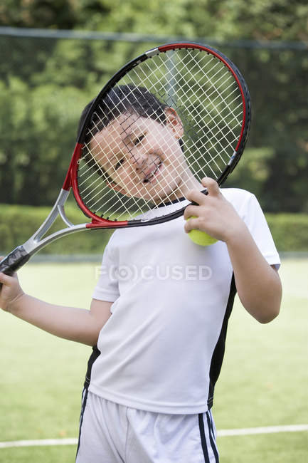 Menino chinês olhando através de raquete no campo de ténis — Fotografia de Stock