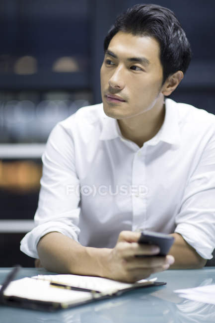 Chinesischer Geschäftsmann hält Smartphone im Büro und schaut weg — Stockfoto