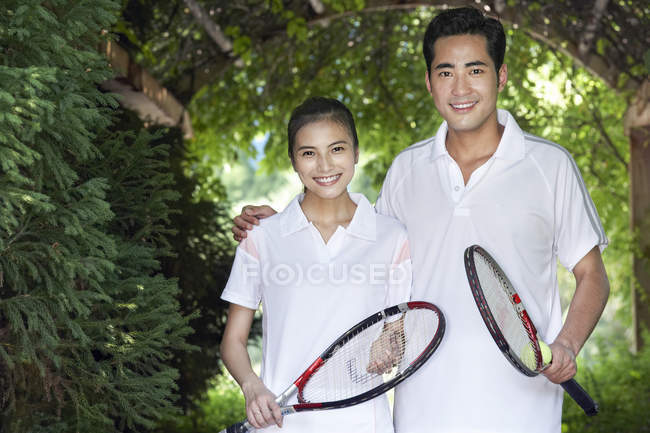 Chinese couple with tennis rackets standing in garden — Stock Photo