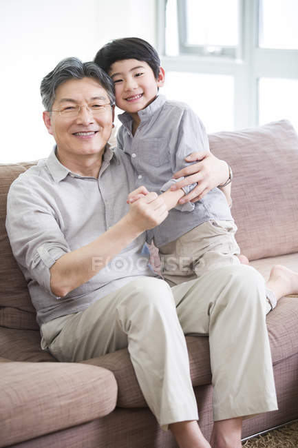 Abuelo y nieto chino abrazándose en el sofá y sonriendo - foto de stock