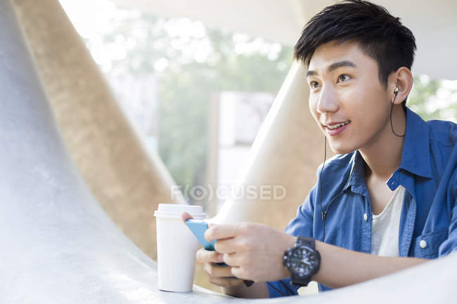 Chinese man listening to music with smartphone and holding coffee — Stock Photo