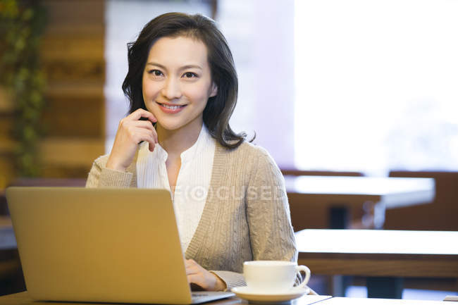 Donna cinese seduta con computer portatile e una tazza di caffè nel caffè — Foto stock