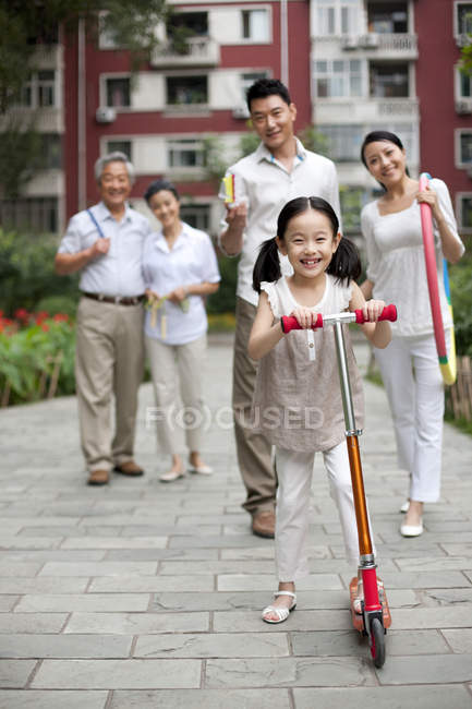 Chica china montando scooter con la familia en el fondo - foto de stock