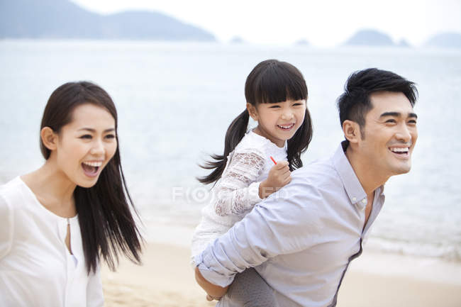 Chinese father giving daughter piggyback ride on beach with mother — Stock Photo
