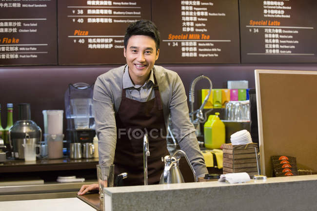 Hombre chino empleado de la tienda de café apoyado en el mostrador en la cafetería - foto de stock
