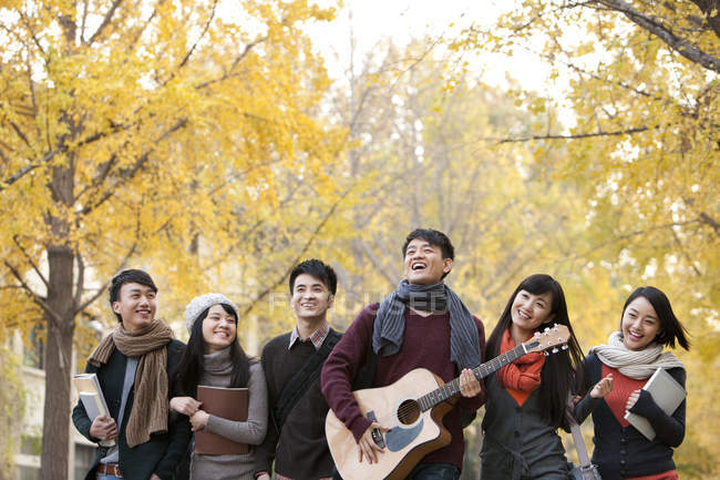 Estudiante universitario chino tocando la guitarra con amigos en el campus en otoño - foto de stock