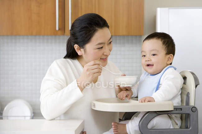 Chinesin füttert Baby-Sohn im Hochstuhl in Küche — Stockfoto