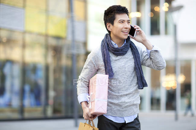 Hombre chino de moda sosteniendo regalo y hablando por teléfono en la calle - foto de stock