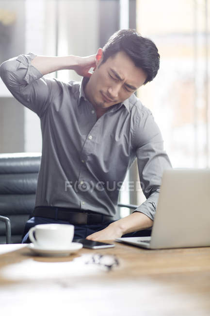 Уставший китаец с болью в шее сидит с ноутбуком в офисе — стоковое фото