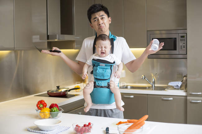 Chinese father holding crying baby and shrugging in kitchen — Stock Photo