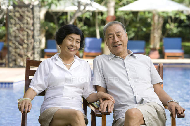 Senior Chinese couple sitting in chairs by hotel pool — Stock Photo