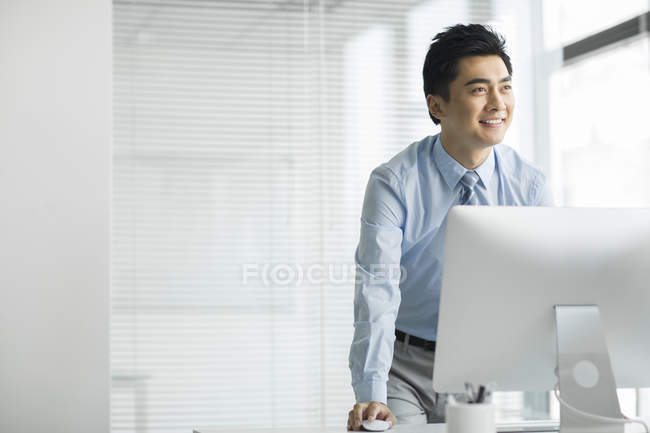 Китайский бизнесмен стоит за компьютером в офисе и смотрит в сторону — стоковое фото