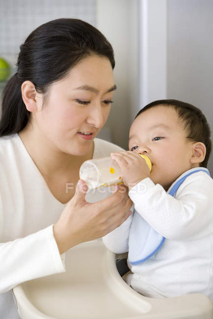 Chinês mulher alimentando bebê filho com garrafa em cadeira alta na cozinha — Fotografia de Stock