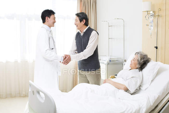 Chinese man shaking hands with doctor with woman in hospital bed — Stock Photo