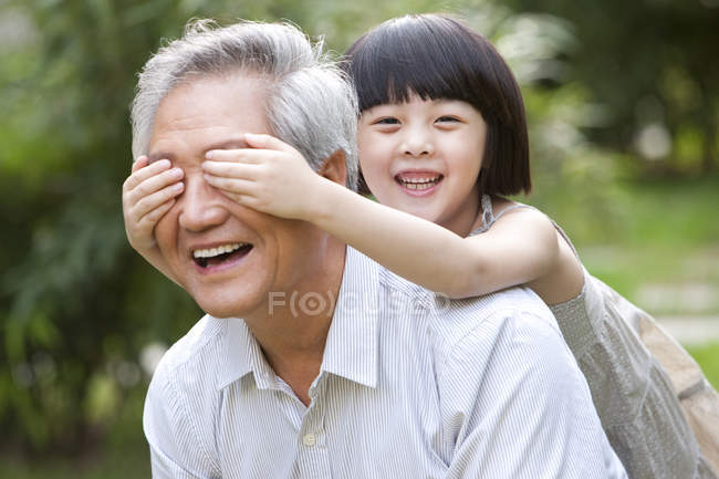 Menina chinesa cobrindo os olhos do avô com as mãos no jardim — Fotografia de Stock