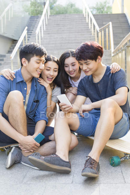 Amigos chinos mirando el teléfono inteligente en las escaleras con patinetas - foto de stock