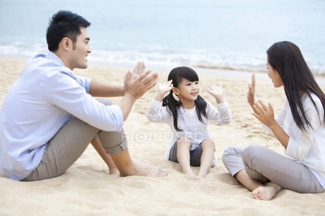 Chinesische Eltern mit Tochter sitzen am Strand und klatschen in die Hände — Stockfoto
