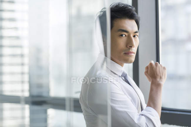 Pensativo empresario chino de pie detrás de la pared de cristal en la oficina - foto de stock