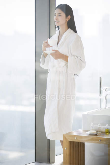 Жінка в халаті тримає каву і дивиться через вікно у ванній — стокове фото