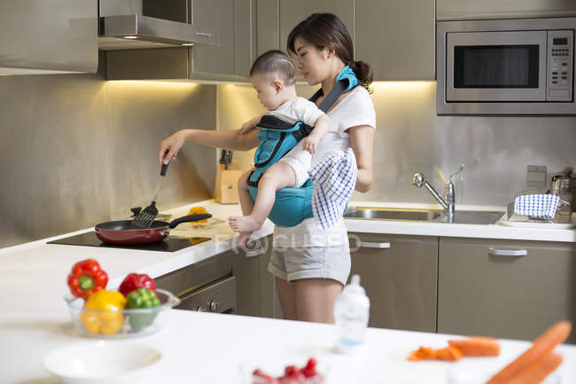 Chinese mother holding baby and cooking in kitchen — Stock Photo