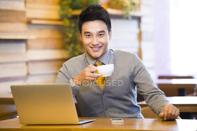 Chinese man drinking coffee in coffee shop with laptop on table — Stock Photo