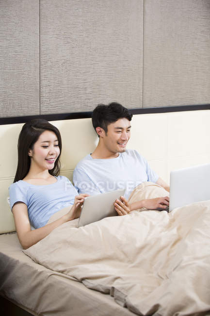 Joven pareja china usando dispositivos inalámbricos en la cama - foto de stock