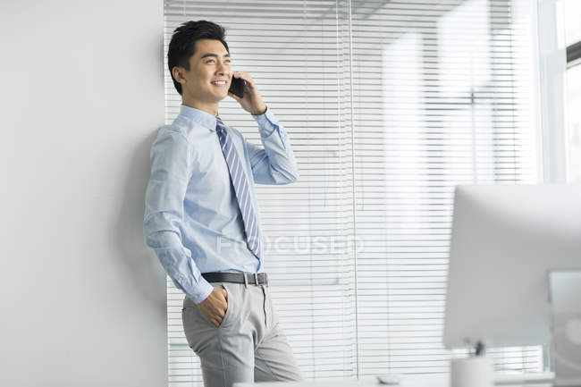 Chinese businessman leaning on wall and talking on phone in office — Stock Photo