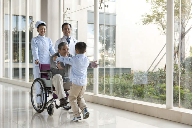 Neto chinês correndo para o avô em cadeira de rodas com médicos — Fotografia de Stock