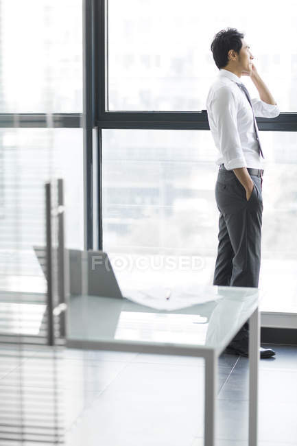 Homme d'affaires chinois parlant au téléphone et regardant par la fenêtre dans le bureau — Photo de stock