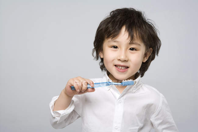 Menino asiático segurando escova de dentes no fundo cinza — Fotografia de Stock